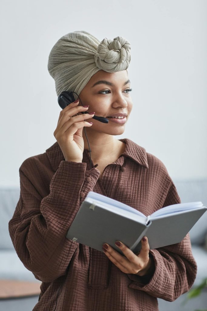 Positive African American female call agent in headset wearing turban standing in light room with notepad in light room at home