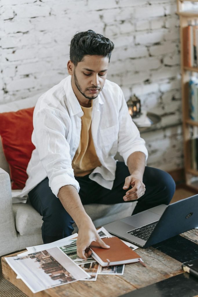 Young Hispanic male designer taking planner from table with laptop and pictures while working on project in modern loft style workplace